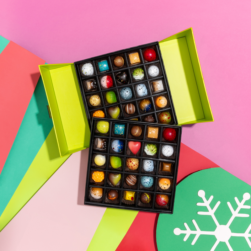 A stunning green tiered gift box featuring all 10 limited-edition holiday chocolates alongside 40 pieces from our signature collection. The gift box is on a pink and multicolored background.